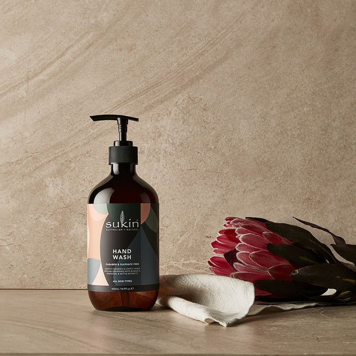 Sukin hand wash bottle on a wooden surface with a pink flower and white towel against a beige wall.