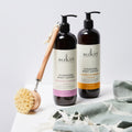 Two bottles of Sukin body lotion with a brush and towel on a light background