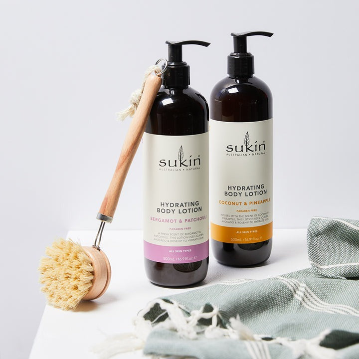 Two bottles of Sukin body lotion with a brush and towel on a light background